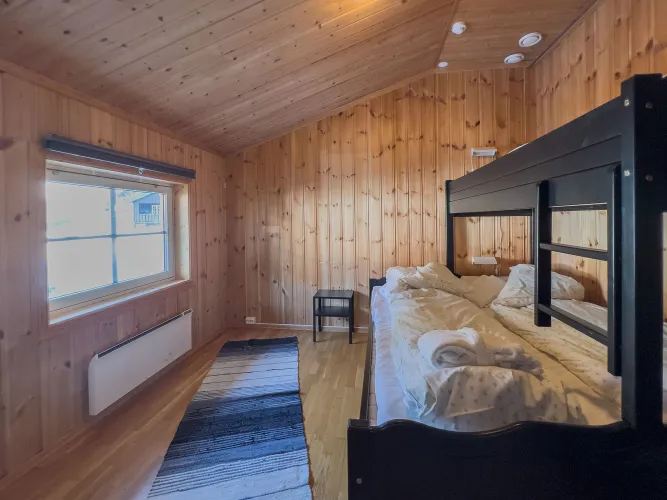 Houten slaapkamer met stapelbed, opgemaakt beddengoed, klein nachtkastje, vloerkleed en raam met daglicht..