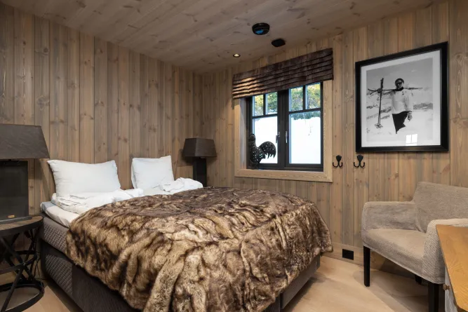Houten slaapkamer met tweepersoonsbed en bontdeken, raam met sneeuwzicht, lampen, fauteuil en ingelijste foto..