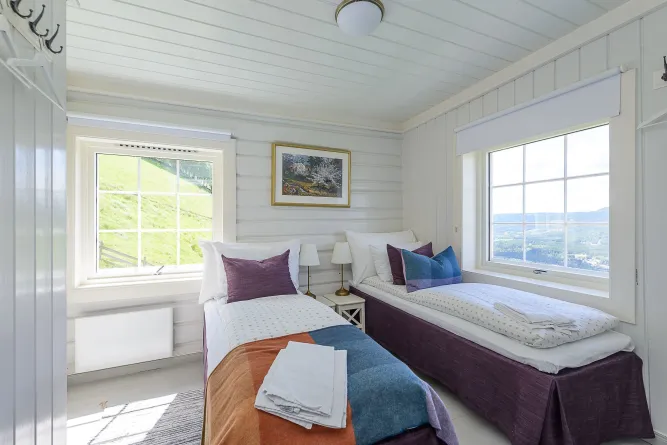 Helles Schlafzimmer mit zwei Einzelbetten, bunten Kissen, Nachttischlampen und zwei Fenstern mit Bergblick..