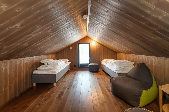 Holzverkleideter Dachbodenraum mit zwei Einzelbetten, dunklem Sitzsack, Hocker und Türfenster am Giebel..
