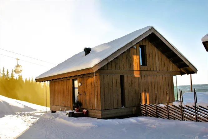 Eine Holzhütte mit schneebedecktem Dach in einer Winterlandschaft, warmes Sonnenlicht und eine Gondel im Hintergrund..