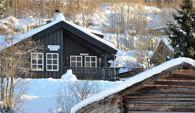 Donkere houten hut met veranda en besneeuwd dak in een winterlandschap met bomen en een houten schuur op de voorgrond..