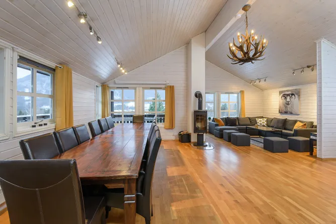 Interieur van een houten chalet met lange eettafel, zwarte stoelen, houtkachel, hoekbank en grote ramen..