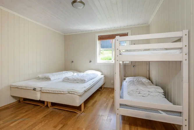 Eenvoudige slaapkamer met een tweepersoonsbed links en een wit stapelbed rechts, met raam en houten vloer..