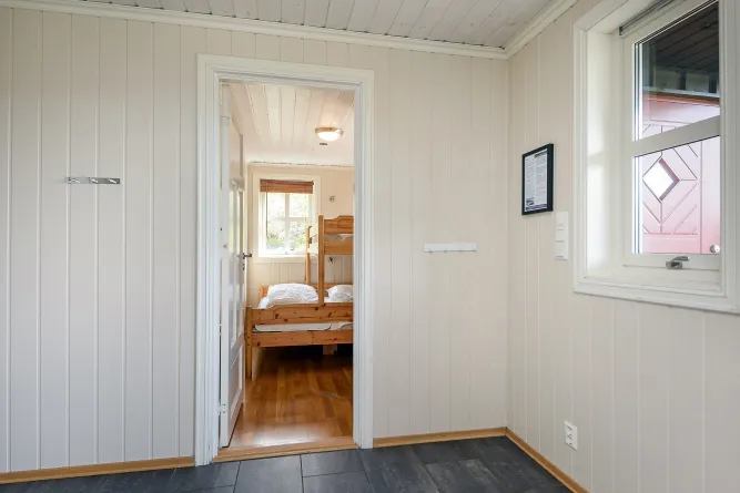 Lichte hal met witte houten wanden en tegelvloer, met doorkijk naar slaapkamer met stapelbed en raam..
