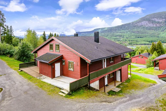 Rotes Holzhaus mit dunklem Dach und Balkon auf grünem Hang, dahinter Waldberge und blauer Himmel mit Wolken..