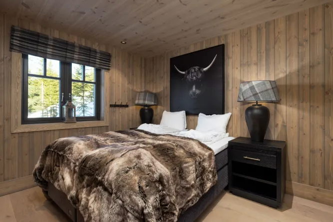 Rustieke houten slaapkamer met tweepersoonsbed, bontplaid, nachtkastje met lampen en raam met boszicht..