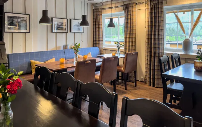 Heller Innenraum eines Restaurants mit Holztischen, Stühlen, Sitzbank, Pendelleuchten, Kerzen und Fenstern mit Vorhängen..