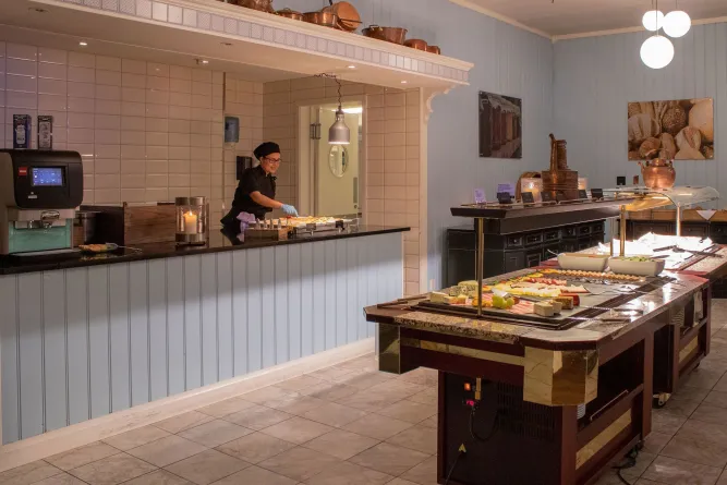 Innenraum mit Buffetinsel mit Käse und Aufschnitt; dahinter bereitet eine Küchenmitarbeiterin Speisen am Tresen zu..