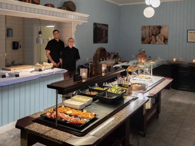 Zwei Köche stehen hinter einem Buffet mit warmen Speisen, Salaten und Platten in einem hellblauen Gastraum..