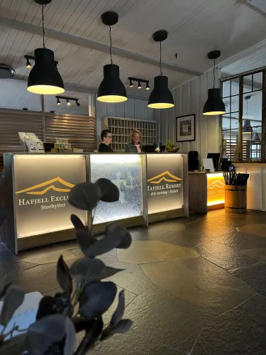 Rezeptionsbereich eines Hotels mit beleuchtetem Tresen, Pendelleuchten, zwei Mitarbeitern und Schlüsselfächern im Hintergrund..