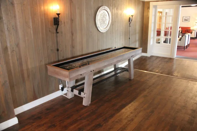 Langer Shuffleboard-Tisch in einem Raum mit Holzvertäfelung, warmen Wandleuchten und Tür zu einem Sitzbereich..