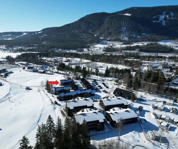 Luchtfoto van besneeuwde gebouwen en een parkeerplaats bij een open sneeuwveld, met beboste bergen op de achtergrond..