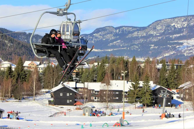 Twee skiërs in een stoeltjeslift boven een besneeuwd skigebied met chalets, bomen en bergen onder blauwe lucht..