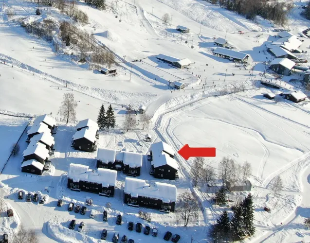 Luchtfoto van besneeuwde skiresortgebouwen en pistes met sporen, kleine figuren en een rode pijl naar een chalet..