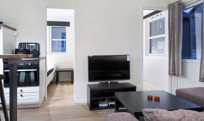 Heldere woonkamer met flatscreen op tv-meubel, salontafel, bank, ramen met gordijnen en kitchenette links..