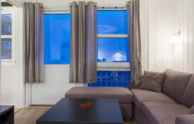 Gezellige woonkamer met grijze hoekbank, zwarte salontafel en grote ramen met grijze gordijnen bij schemerlicht..