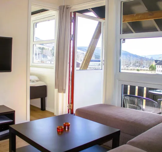 Interieur met bank, salontafel en open balkondeur; door grote ramen zicht op bergen en een dorpslandschap..