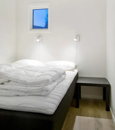 Minimalistische slaapkamer met tweepersoonsbed, wit dekbed en kussens, twee wandlampjes, klein raam en nachtkastje..