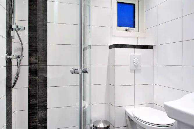 Witte betegelde badkamer met glazen douchecabine, chrome douchekraan, toilet en klein raam met blauw glas..