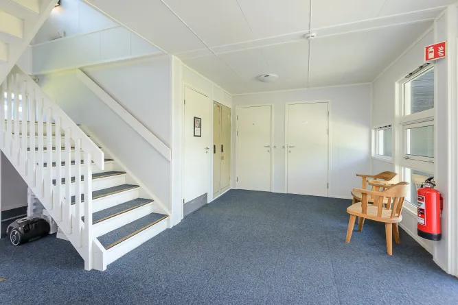 Lichte entreehal met witte trap links, twee deuren achterin, ramen rechts, houten stoelen en brandblusser..