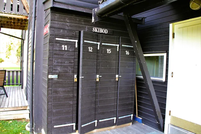Zwarte houten skiberging met vier deuren, nummers 11, 12, 15 en 16, hangsloten en het bordje 'SKIBOD'..