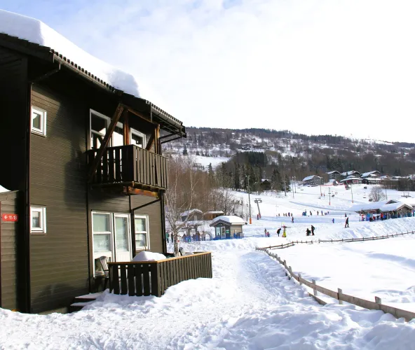 Donker houten chalet met balkon naast een besneeuwd pad, met skiërs en pistes op de heuvels in de verte..