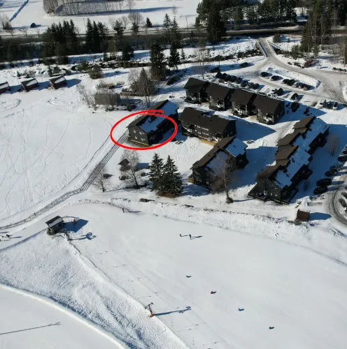 Luchtfoto van een besneeuwd vakantiepark met donkere chalets, parkeerplaats en een gebouw in rode cirkel..