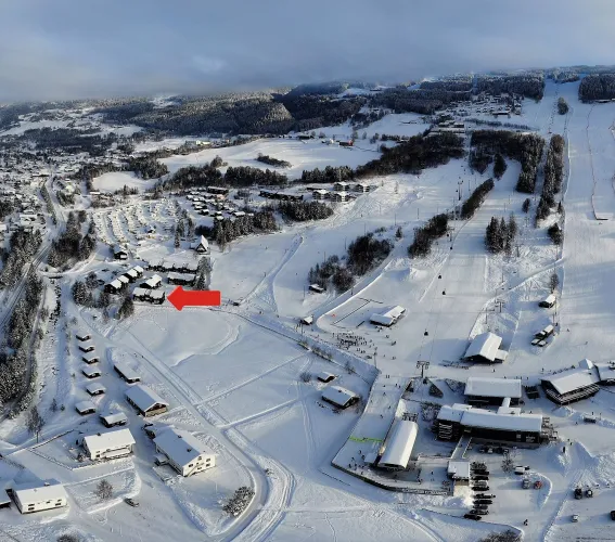 Luchtfoto van een besneeuwd skigebied met skipistes, chaletdorp en gebouwen; een rode pijl wijst een locatie aan..