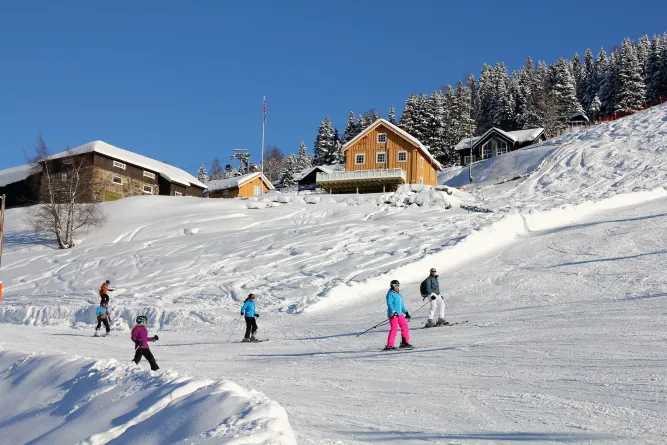 Mehrere Skifahrer auf einer verschneiten Piste; darüber stehen Berghütten und ein verschneiter Wald unter blauem Himmel..