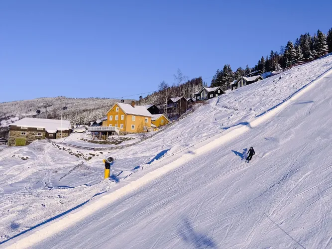 Ein Skifahrer fährt eine verschneite Skipiste hinab; im Hintergrund stehen Hütten am Hang unter blauem Himmel..