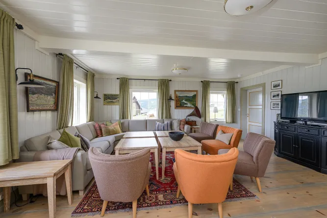 Helles Wohnzimmer mit großem Ecksofa, Sesseln, Holztischen, Teppich, Bildern an der Wand und Fernseher im Eck..