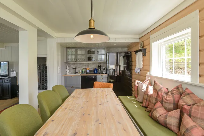 Helle Wohnküche mit langem Holztisch, grüner Bank mit Kissen, grauen Schränken und großem Fenster rechts..