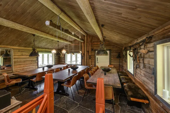 Großer rustikaler Speiseraum mit langen Holztischen, braunen Stühlen, Hängelampen und Holzdecke in einer Blockhütte..