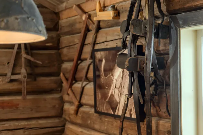 Innenansicht einer Blockhütte mit hängendem Pferdegeschirr, Werkzeugen an der Wand, Lampe und gerahmtem Bild..
