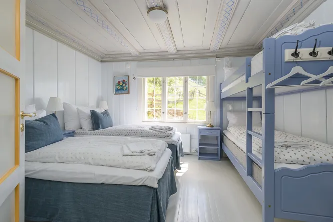 Heller Schlafraum mit Doppelbett und blauem Etagenbett, weißen Wänden, Nachttischen, Lampen und Fensterlicht..