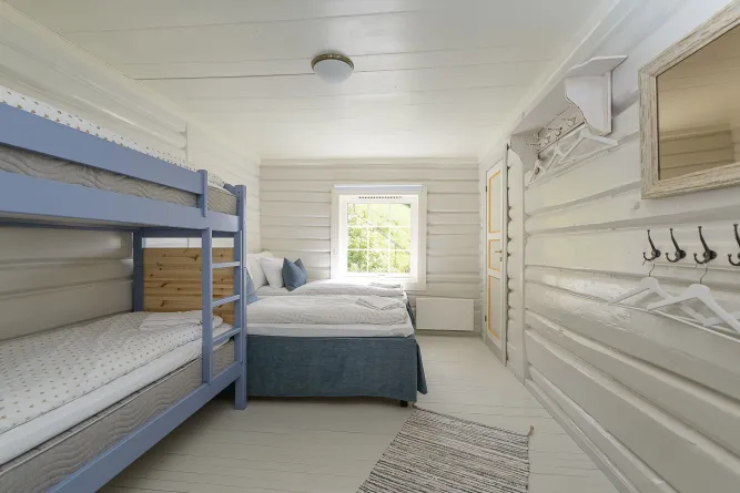 Helles Schlafzimmer mit blauem Etagenbett, Doppelbett, Fensterblick ins Grüne, weißer Holzvertäfelung und Kleiderhaken..
