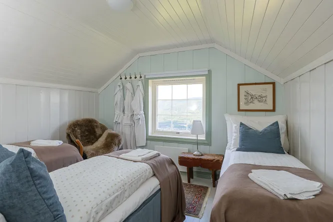 Helles Dachzimmer mit zwei Einzelbetten, Fensterblick, Bademänteln an Haken, kleinem Tisch mit Lampe und Bild an der Wand..