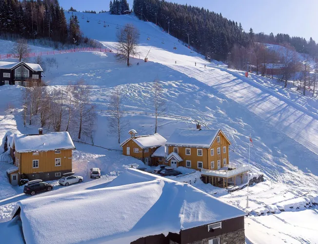 Verschneite Holzhäuser am Fuß eines Skihangs mit Sessellift, Bäumen und wenigen Skifahrern im Sonnenschein..