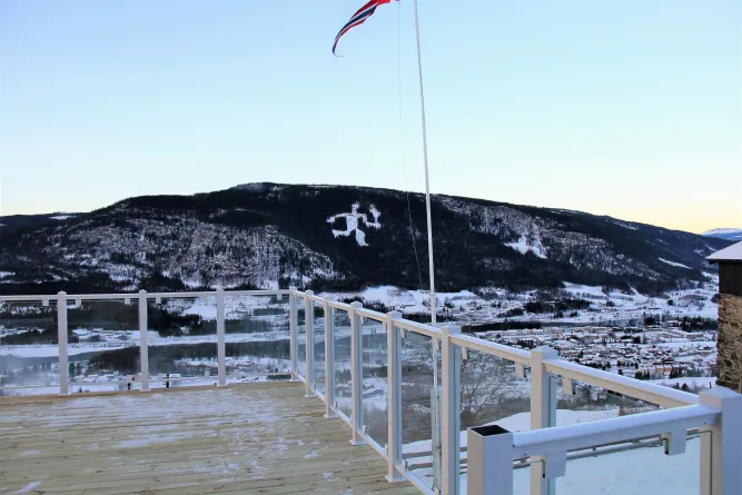 Holzterrasse mit Glasgeländer und Fahnenmast, Blick auf verschneites Tal und bewaldeten Berg unter klarem Himmel..