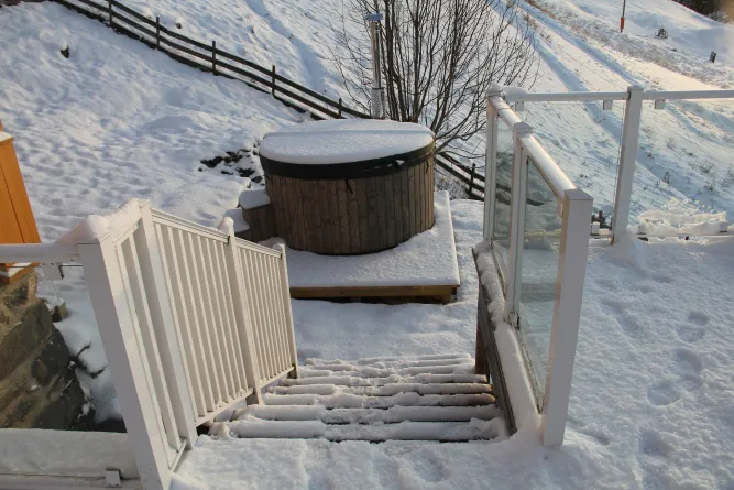 Verschneite Treppe führt zu einem runden Holz-Whirlpool auf einer Terrasse, umgeben von Geländern und Hang im Winter..