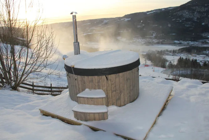 Verschneiter runder Hot Tub aus Holz mit Schornstein auf Terrasse; Dampf steigt auf, Berge und Tal im Hintergrund..