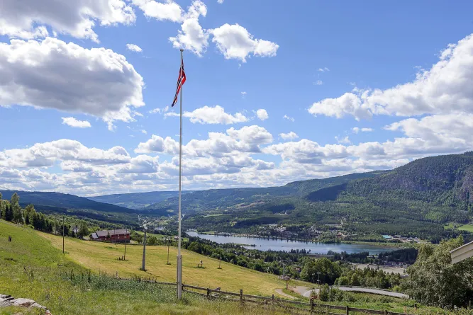 Blick über eine grüne Hangwiese mit Flaggenmast auf ein Tal mit See, Wäldern und Bergen unter Wolkenhimmel..