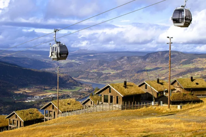Gondeln einer Seilbahn schweben über einer Hangwiese mit Holzhäusern; dahinter ein weites Tal und Wolken..