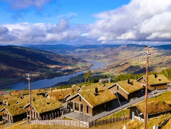 Holzhäuser mit Grasdächern am Hang, Blick über ein breites Flusstal mit Wasserlauf, Bergen und Wolkenhimmel..