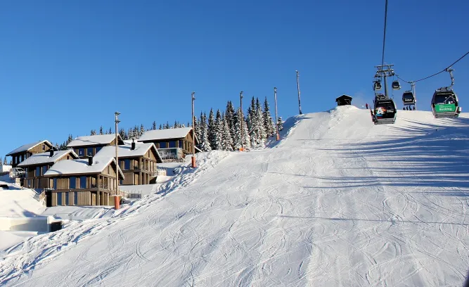 Verschneite Skipiste bei Sonnenschein mit Gondelbahn rechts, Chalets links und Tannenwald am Hang unter blauem Himmel..