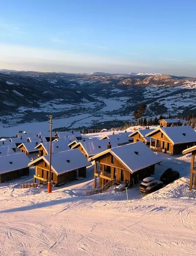 Verschneite Holzhütten an einem Hang mit Skispuren, Blick auf ein weites Tal und Berge unter blauem Himmel..