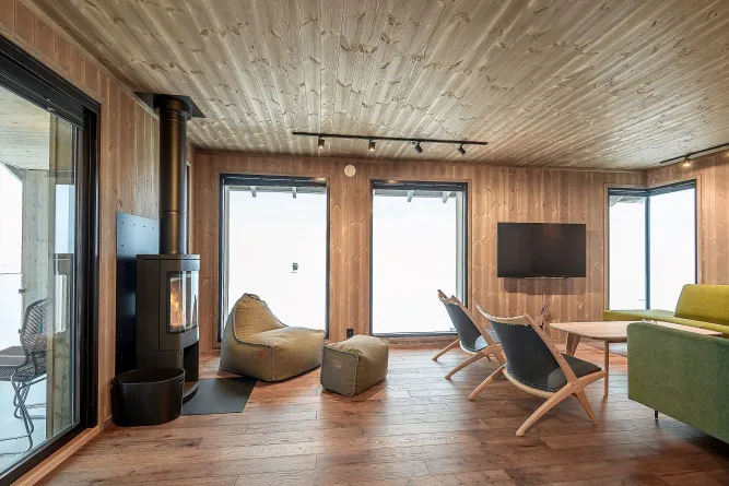 Holzvertäfeltes Wohnzimmer mit Kaminofen, Sitzsäcken, Sesseln, Couchtisch, Sofa und großen Fenstern mit Ausblick..