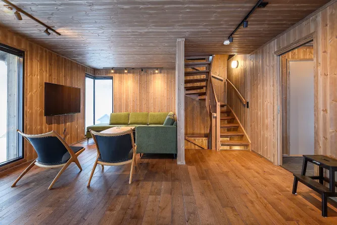 Helles Wohnzimmer mit Holzvertäfelung, grüner Eckcouch, zwei Sesseln, Wand-TV und Treppe nach oben..