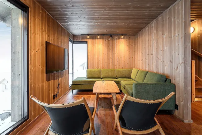 Moderner Wohnraum mit Holzvertäfelung, grünem Ecksofa, Couchtisch, zwei Sesseln und TV an der Wand..